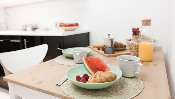 A breakfast setting with a plate of croissants, watermelon, and raspberries, a coffee cup, and a bottle of orange juice on a wooden table. - Warsaw Concierge Square Grzybowski (Warsaw)