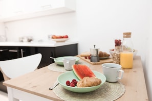 Desayuno con un plato de cruasanes, sandía y frambuesas, una taza de café y una botella de jugo de naranja sobre una mesa de madera.