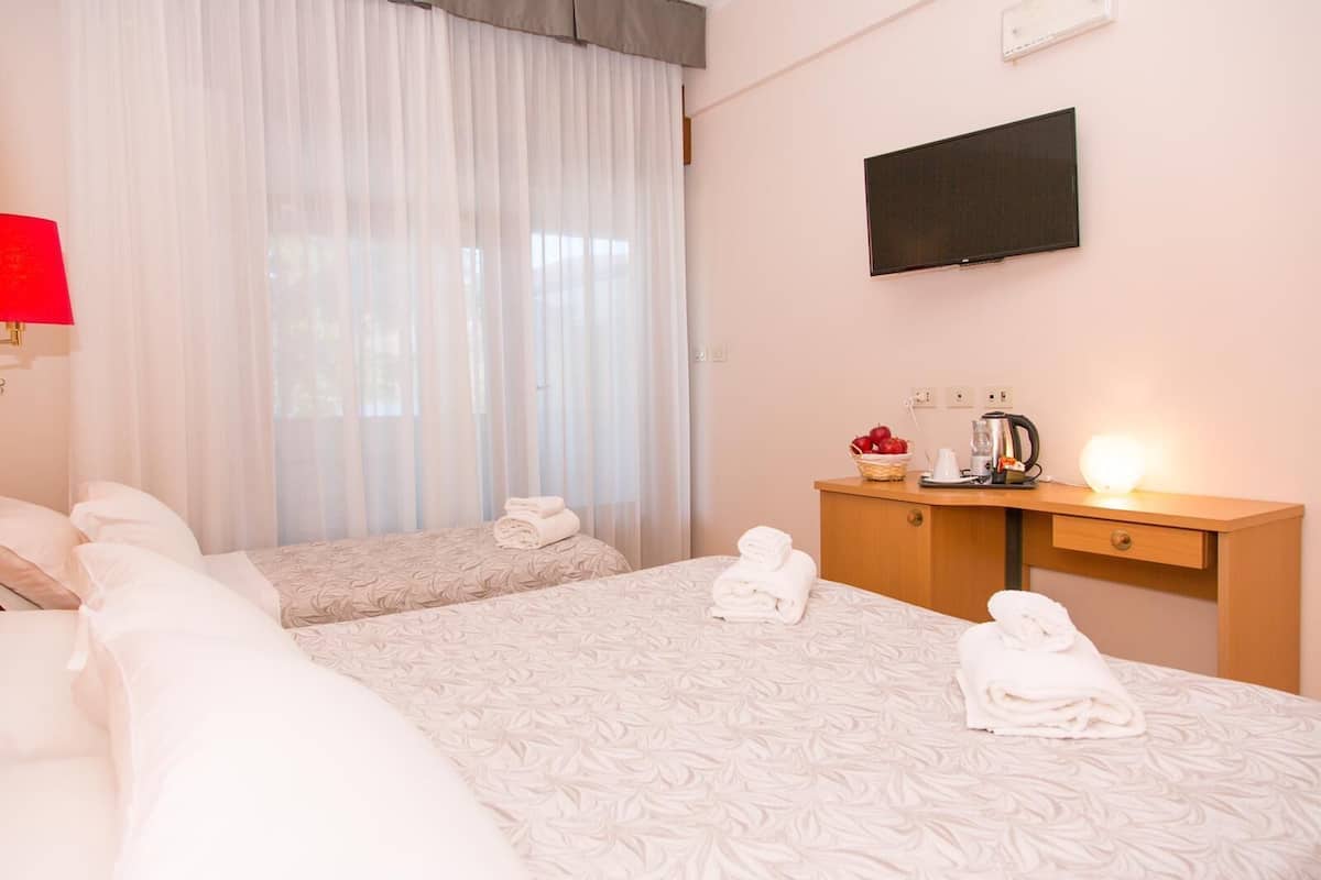 A hotel room with two beds, a desk with a TV, and a window with sheer curtains.