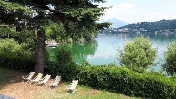 A lakeside view with a row of sun loungers under a tree, a clear sky, and a mountain in the background.
