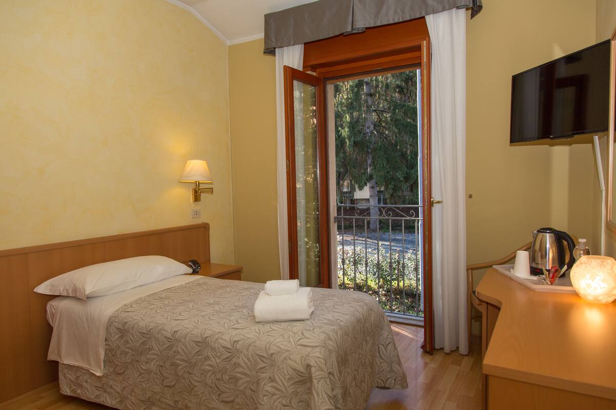 A hotel room with a bed, a nightstand, a desk with a kettle, a flat-screen TV, and a window with a view of greenery.