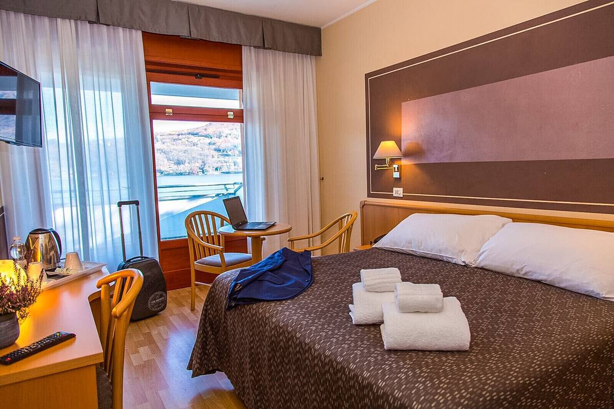 A hotel room with a bed, a desk with a laptop, a chair, a TV, and a view of a body of water through the window.