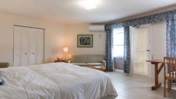 A hotel room with a large bed, a desk, a chair, a nightstand, a lamp, a window with curtains, and a door. - Clairfont, Jobson's Cove / Warwick Long Bay/ Horseshoe Beach -  APARTMENT 2 (Warwick)