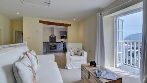 Un salón moderno con un sofá, un sillón y una mesa de centro de madera con revistas. Se ve una cocina al fondo, y un balcón con vistas.