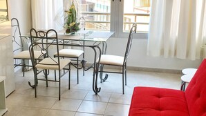 Spiseområde med et glasbord og metalstole, en rød sofa og udsigt til den udendørs balkon.
