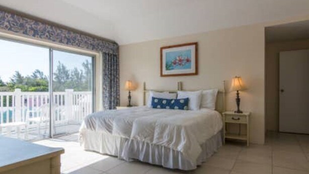 A bedroom with a large bed, two bedside tables with lamps, a window with a view of trees, and a door. - Clairfont, Jobson's Cove / Warwick Long Bay/ Horseshoe Beach -  APARTMENT 6 (Warwick)