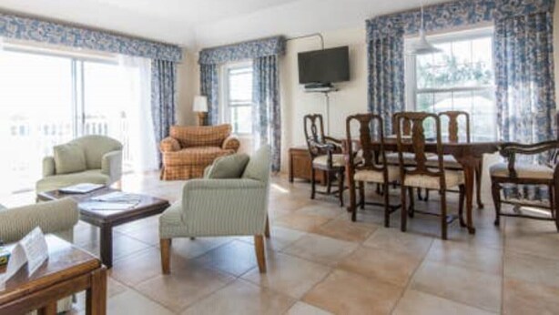A spacious living room with a sofa, armchairs, a dining table with chairs, a flat-screen TV mounted on the wall, and large windows with curtains. - Clairfont, Jobson's Cove / Warwick Long Bay/ Horseshoe Beach -  APARTMENT 8 (Warwick)