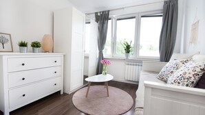 A modern, well-lit room with a white dresser, a round table with a vase of flowers, a sofa with patterned cushions, and a window with curtains.