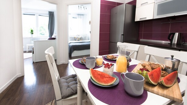 A modern kitchen with a dining table set for breakfast, including watermelon, juice, and pastries.