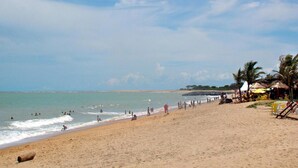 A beach with people swimming and walking along the shore, palm trees, and beach umbrellas.
