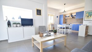 A modern living room with a TV, a dining table, and a kitchen area with blue cabinets. - Sitting on the beach (Santa Croce Camerina)