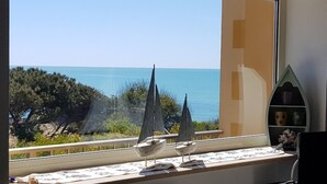 A room with a large window overlooking a coastal view, featuring a small shelf with decorative items. - Sitting on the beach (Santa Croce Camerina)