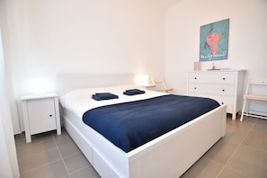 A bedroom with a white bed, a nightstand, a dresser, and a wall-mounted artwork.