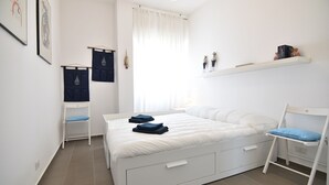 A bedroom with a bed, a chair, a shelf, and a window. - Sitting on the beach (Santa Croce Camerina)