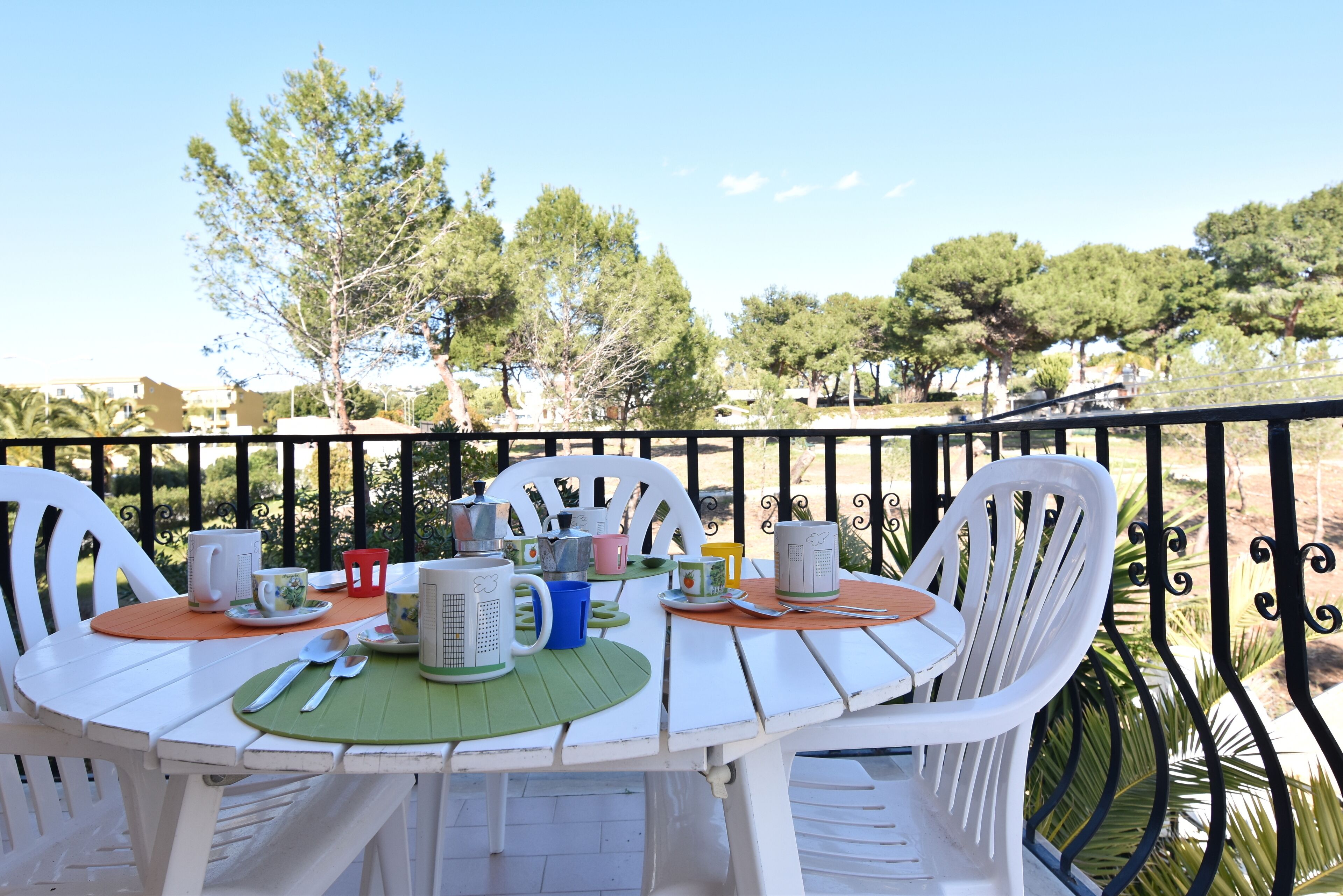 Una mesa al aire libre con sillas blancas, un mantel individual verde y varias tazas y vasos.