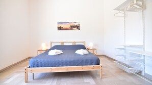 A neatly made bed with a blue quilt, two bedside lamps, and a cityscape painting on the wall. - Center Beach (Ragusa)