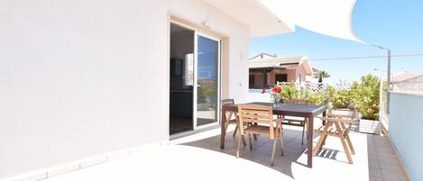 Un patio moderno al aire libre con puerta corrediza que da a un balcón, una mesa de madera con sillas y una vela de sombra blanca.