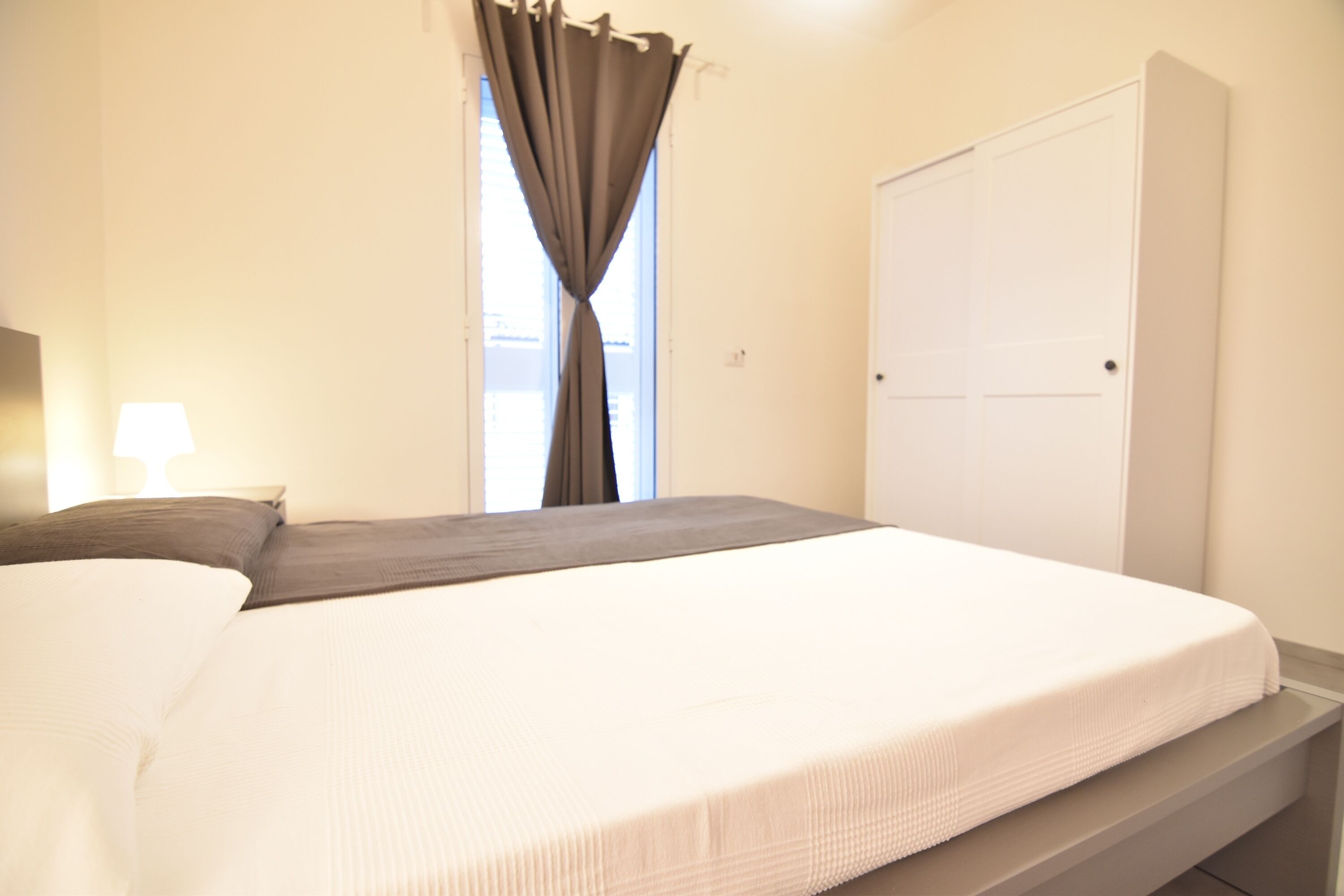 A neatly made bed with a white and gray bedspread, a dark gray pillow, and a white pillow. There is a window with a curtain, a white door, and a wall-mounted light fixture.