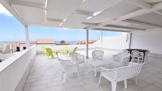 Un patio techado al aire libre con mobiliario de plástico blanco y verde, piso de baldosas y vista al mar y edificios al fondo.