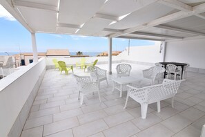 A covered outdoor patio with white and green plastic furniture, a tiled floor, and a view of the sea and buildings in the background. - Casa del mar (Ragusa)