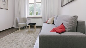 Un salon avec un canapé gris, un fauteuil blanc, une petite table et une fenêtre avec des rideaux.