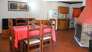 A dining area with a red tablecloth, wooden chairs, a kitchen with a brick fireplace, and a tiled floor.