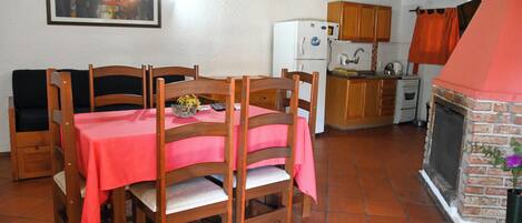 Un comedor con mantel rojo, sillas de madera, una cocina con chimenea de ladrillo y piso de baldosas.