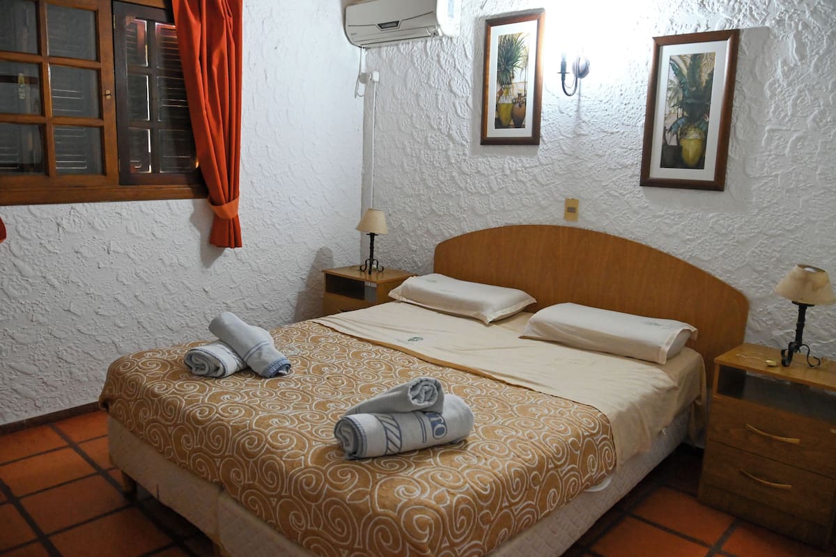 A hotel room with a bed, a nightstand, a lamp, a window with curtains, and a wall-mounted air conditioner.