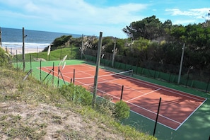 Een tennisbaan met een groen en rood oppervlak, omringd door een hek, met zicht op de oceaan en bomen op de achtergrond.