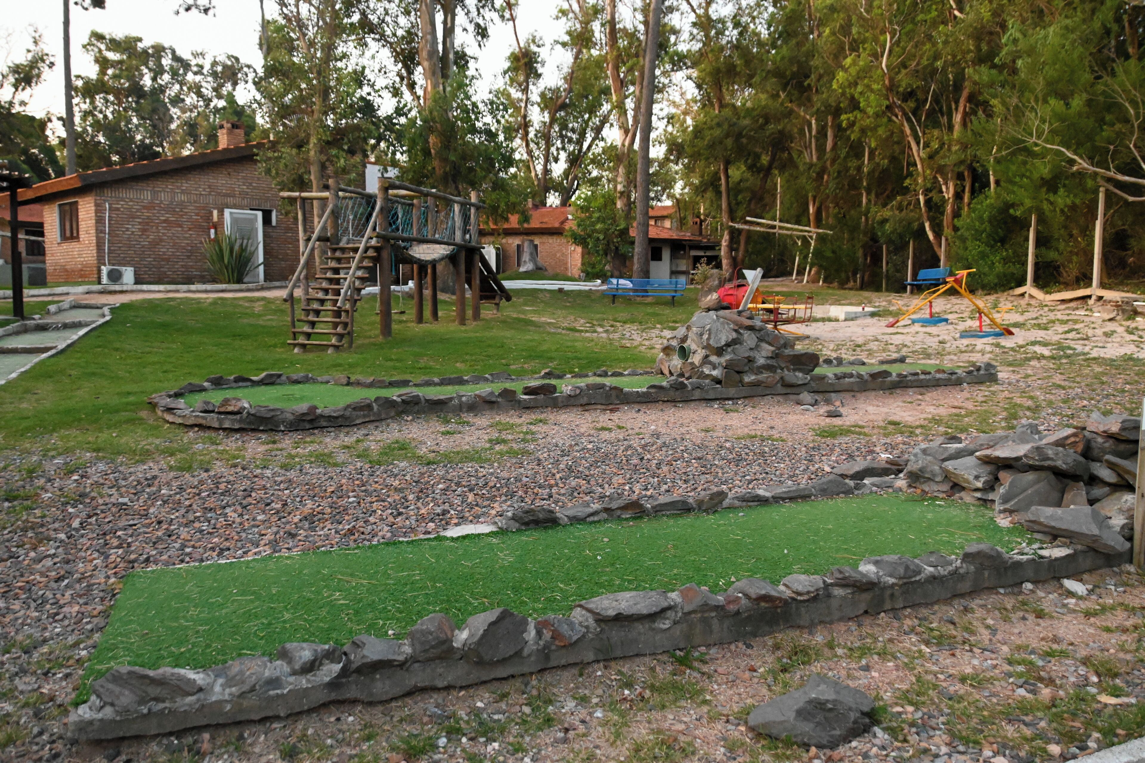 A playground with a climbing structure, a small building, and a grassy area with a rock border.