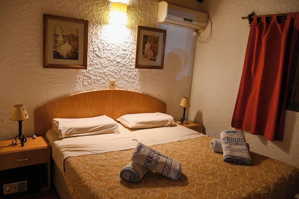 A hotel room with a bed, two wall-mounted lamps, a nightstand, a red curtain, and two framed pictures on the wall.
