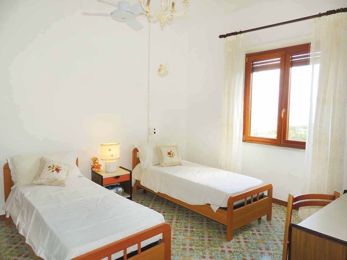 Une chambre avec deux lits simples, une table de chevet avec une lampe, une chaise en bois et une fenêtre avec des rideaux.