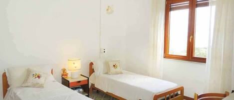 Une chambre avec deux lits simples, une table de chevet avec une lampe, une chaise en bois et une fenêtre avec des rideaux.