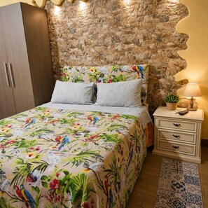 Une chambre avec un mur en pierre, un lit recouvert d’une couvre-lit à motifs floraux, une table de chevet avec une lampe et une armoire en bois.