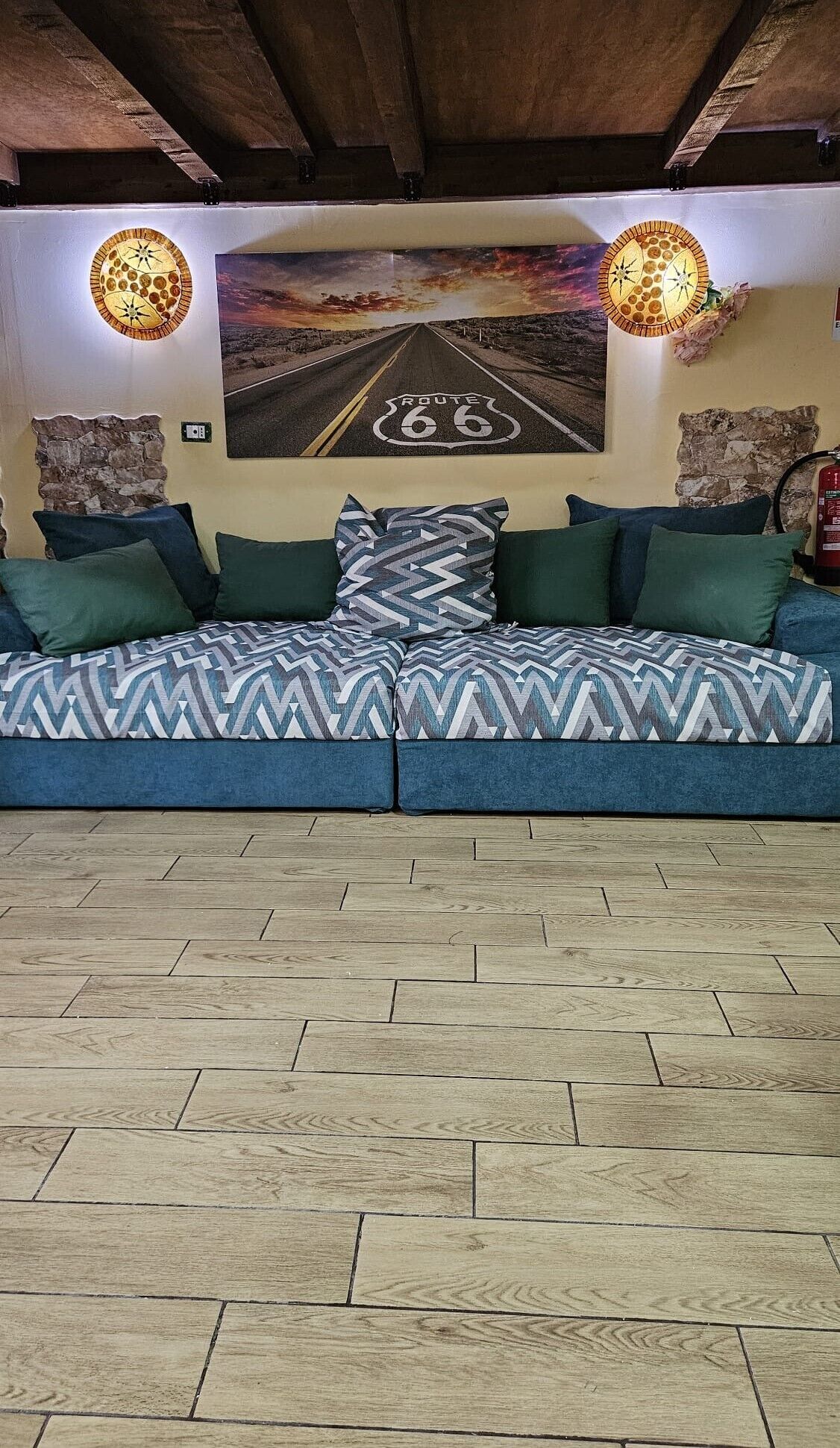 A room with a sectional sofa, a Route 66 road painting, and two wall-mounted lamps.