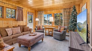 Ein gemütliches Blockhaus-Interieur mit Holzbalkendecke, einer Couch, zwei Sesseln, einem kleinen Tisch und einem Weihnachtsbaum.