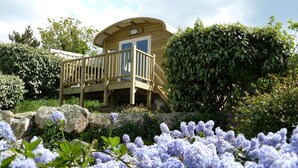 A wooden cabin with a deck, surrounded by lush greenery and purple flowers. - Camping de l'Iroise (Plomodiern)