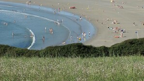 A beach with people swimming and sunbathing, houses in the background, and a grassy foreground. - Camping de l'Iroise (Plomodiern)