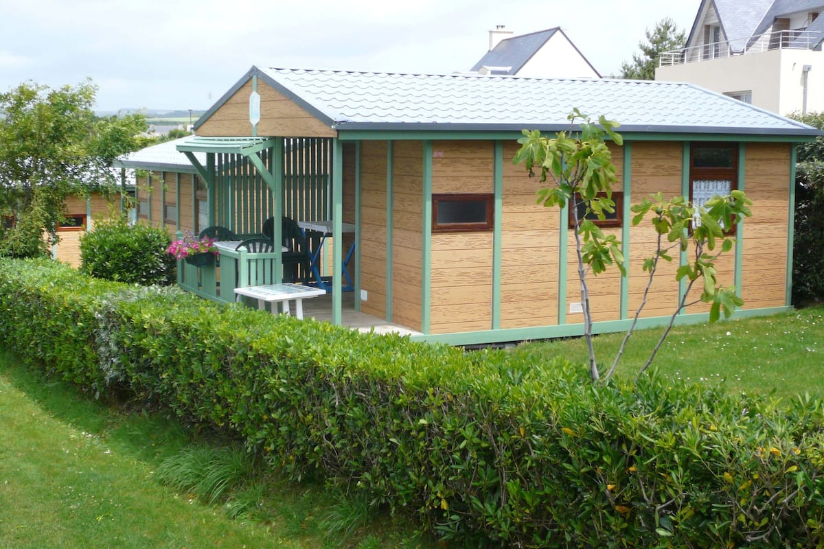 A small, single-story house with a sloped roof, a porch with a green railing, and a garden in front.