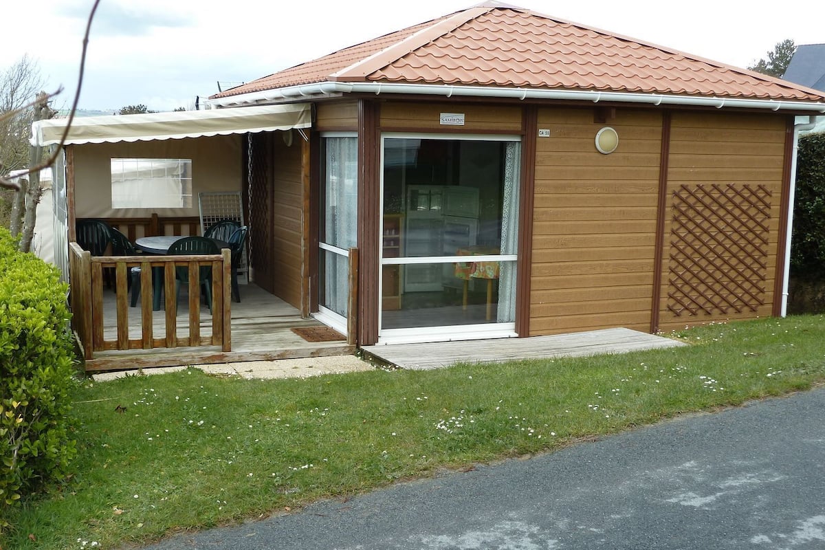 A single-story wooden house with a tiled roof, a covered porch with chairs, and a glass door entrance.