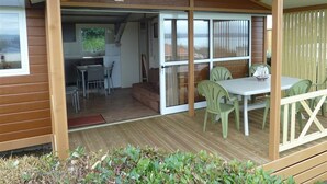 A covered outdoor area with a wooden deck, a table with chairs, and a view of the interior through a glass door. - Camping de l'Iroise (Plomodiern)