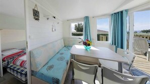 A room with a bed, a table with chairs, a window with curtains, and a view of the outdoors. - Camping de l'Iroise (Plomodiern)