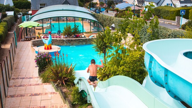 A pool area with a slide, surrounded by greenery and houses. - Camping de l'Iroise (Plomodiern)