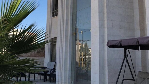 Un edificio con amplias columnas y un puerta de vidrio. Se ve una palmera y mobiliario exterior.