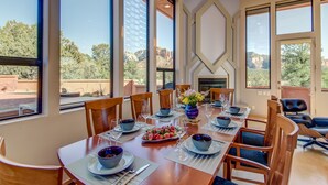 A dining area with a long table set for a meal, large windows offering a view of the outdoors, and a fireplace in the background. - Experience luxury in Sedona Painted Sky—sleeps 8 with stunning panoramic views. (Sedona)