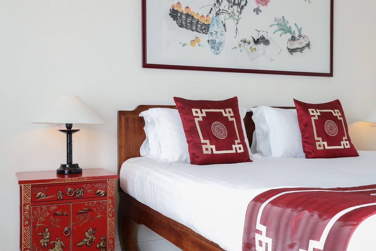 A hotel room with a wooden bed, red decorative chest, and a framed artwork on the wall.