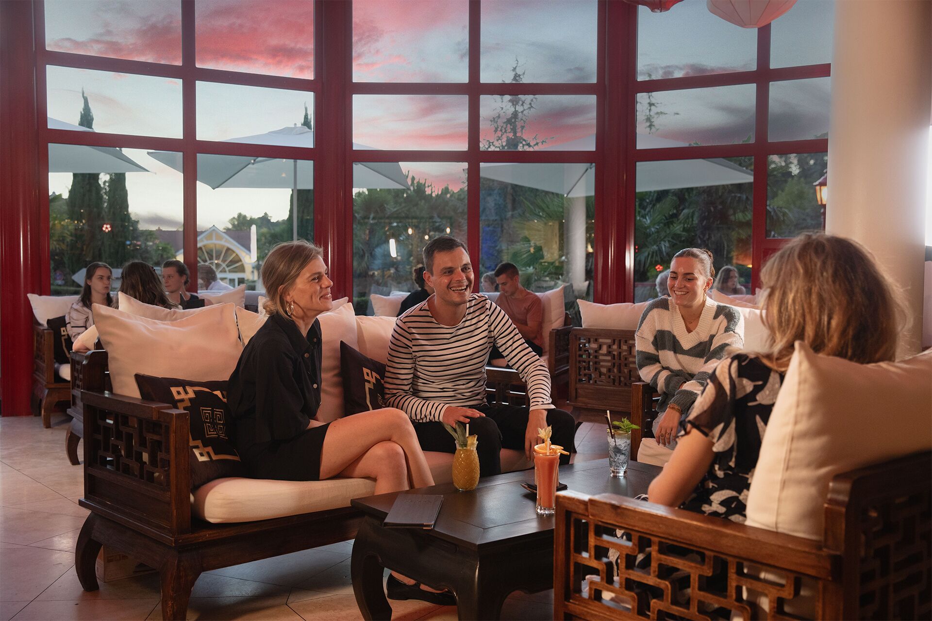 A group of people seated on sofas and chairs in a room with large windows, some with a view of trees and a building.