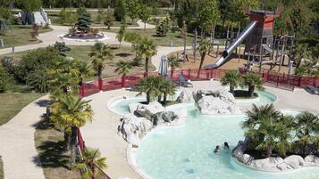 Una piscina con tobogán, rodeada de palmeras y rocas, con valla roja y un parque infantil al fondo.