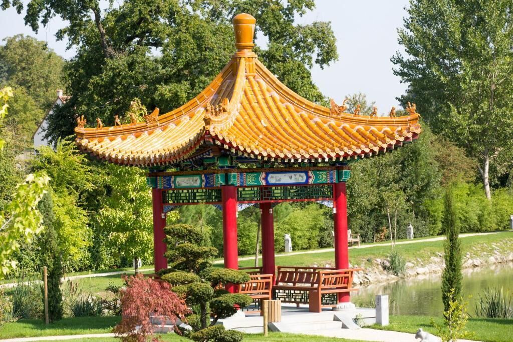 Een klassiek paviljoen met een gouden dak en rode zuilen, omringd door een verzorgde tuin met een vijver en verschillende planten.