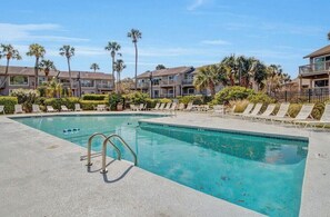 A swimming pool with lounge chairs, surrounded by palm trees and apartment buildings.